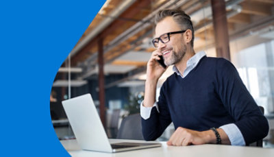 Työpöydän ääressä istuva mies, joka puhuu puhelua matkapuhelimessa ja työskentelee kannettavalla tietokoneella.