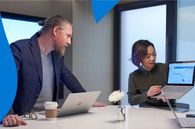 Two people in a meeting room where one person is holding up a laptop to show a coworker information being displayed on the screen.