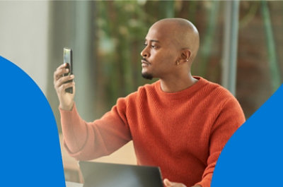 A person sitting down and holding a mobile phone at eye-level while their laptop rests on the table in front of them.