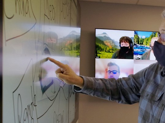 A man wearing a mask is pointing at a whiteboard.