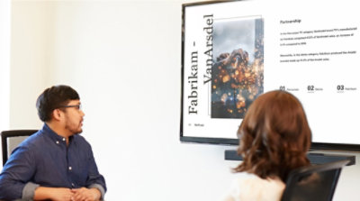 Zwei Personen in einem Konferenzraum, die auf einem großen Display eine Bildschirmpräsentation in PowerPoint ansehen
