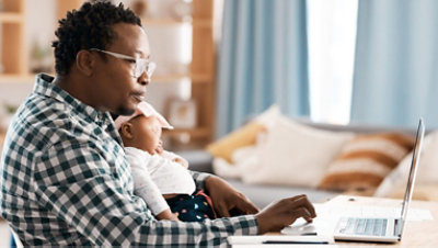 A person wearing glasses uses a laptop while a child sleeps in his lap