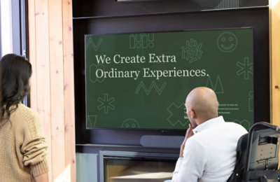 Zwei Personen in einem Besprechungsraum, die eine PowerPoint-Präsentation mit dem Titel „We Create Extra Ordinary Experiences“ ansehen.