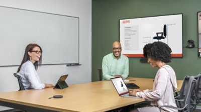 Drei Personen in einem Konferenzraum verwenden Tablets und Laptops, und auf einem großen Bildschirm an der Wand wird eine Präsentation gezeigt