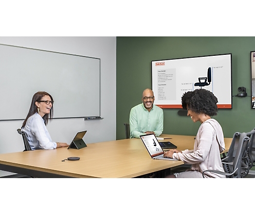 Tiga orang di ruang konferensi menggunakan tablet dan laptop dengan presentasi ditampilkan pada layar besar di dinding