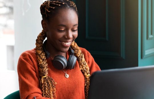 Eine lächelnde Person, die Kopfhörer um den Hals trägt und einen Laptop nutzt