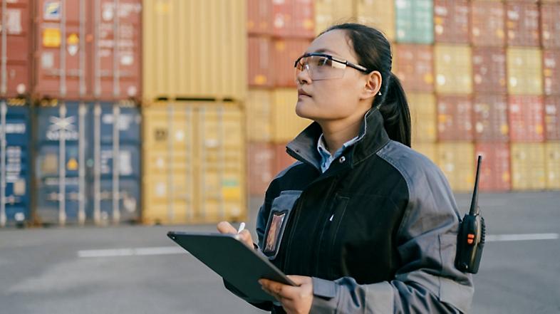 A person wearing protective eyewear standing in front of shipping containers and using a tablet