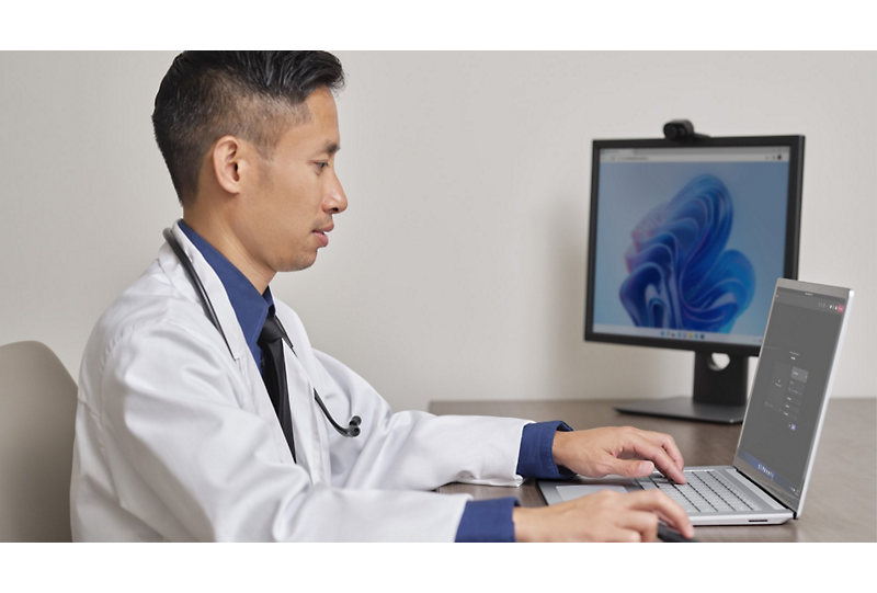 A medical professional wearing a white coat and stethoscope sitting at a desk using a laptop connected to a desktop monitor