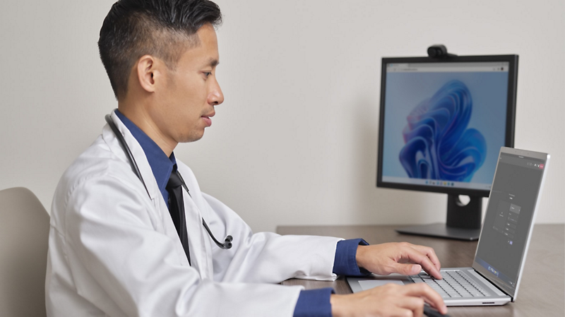 A medical professional wearing a white coat and stethoscope sitting at a desk using a laptop connected to a desktop monitor