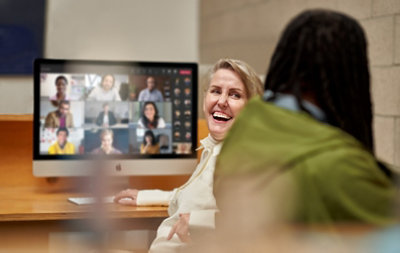 Two people having a conversation while a Teams video call is being displayed on a desktop monitor behind them.