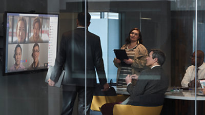 Štiri osebe v konferenčni sobi sodelujejo na sestanku v aplikaciji Microsoft Teams z večjo skupino, ki je prikazana na velikem stenskem zaslonu