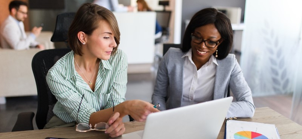 Дві жінки, біла та афроамериканка, співпрацюють за ноутбуком у сучасному офісі.