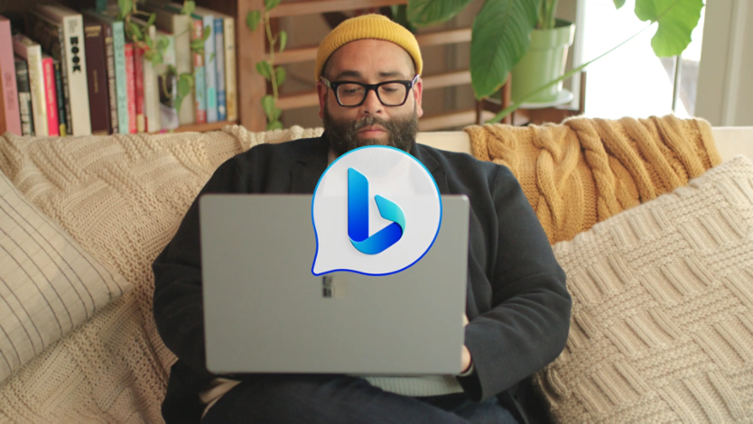 A person holding a computer on their lap while sitting on a couch. In the center is the Bing Chat icon which is a white chat bubble with a blue Bee in the center