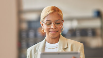 A professional wearing clear-framed glasses looks down at a tablet in an office.