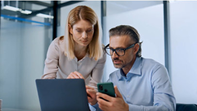 Two coworkers review information on a laptop and smartphone during a discussion.