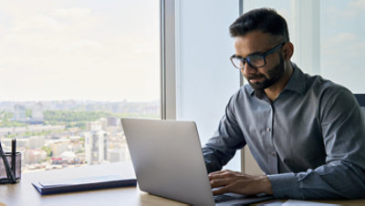 A professional works on a laptop beside a large window overlooking a city skyline.