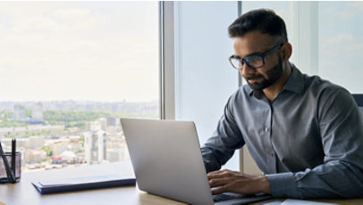 A professional works on a laptop beside a large window overlooking a city skyline.