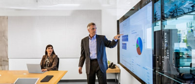 Un uomo che indossa un completo presenta dati su un ampio schermo in una sala conferenze mentre una donna 