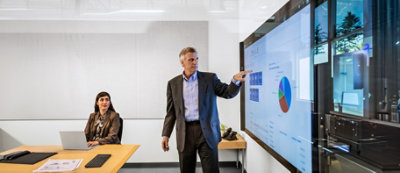 A man in a suit presents data on a large screen to a woman seated at a desk in a modern office.