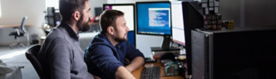Two people sharing a desk and looking at multiple large desktop monitors.