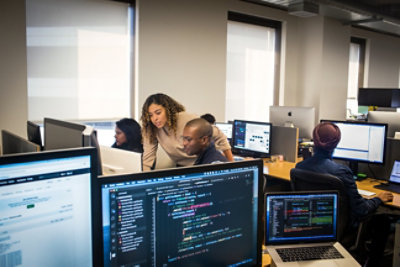Office workspace with people reviewing software and code on multiple computer screens.