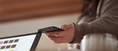 Primo piano di una mano che sorregge uno smartphone vicino allo schermo del portatile, suggerisce 