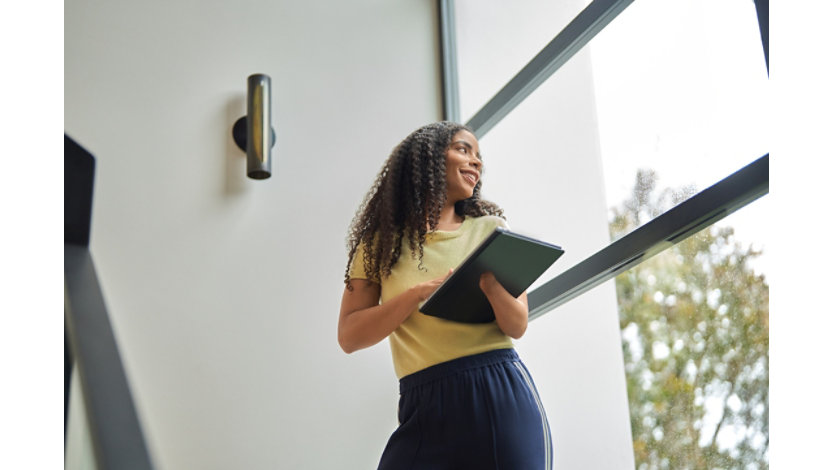 A young professional with a physical disability stands by a window in an office stairwell holding a laptop.
