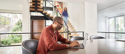 A man sitting at a table using a laptop, with a modern staircase and a blurred person in the background.