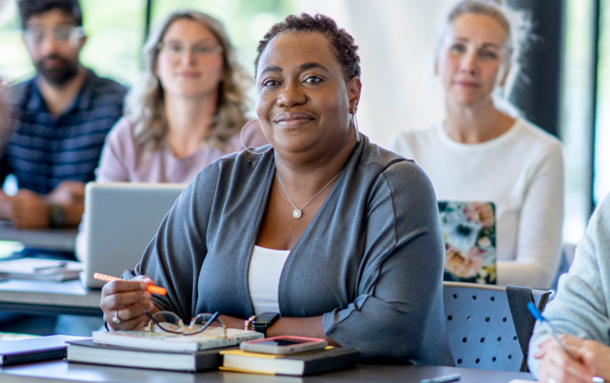 Adult woman attentively sitting in a classroom surrounded by students on laptops.