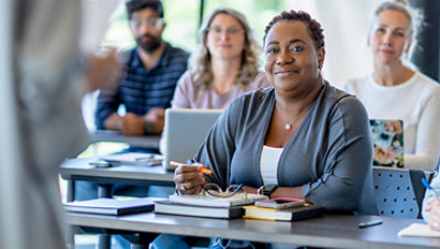 Adult woman attentively listening to a instructor in a classroom surrounded by adult students on laptops.