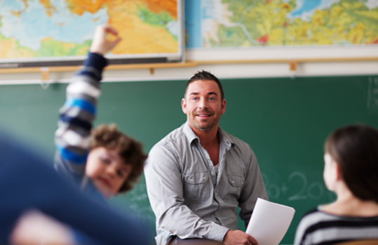 Teacher sits at the front of a class of young students with a boy’s hand raised