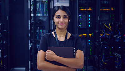 Female IT worker standing in a server room holding a folder and smiling.
