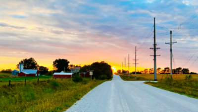 Rural road at sunset with power lines, farm buildings, fields, and trees under a colorful sky.