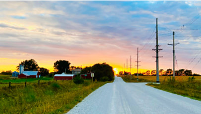 Rural road at sunset with power lines, farm buildings, fields, and trees under a colorful sky.