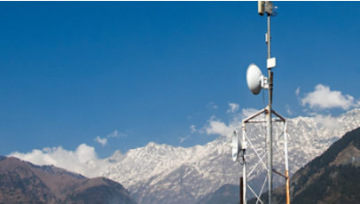 Telecommunications tower with satellite dishes standing against a backdrop of snow-covered mountains under a clear blue sky.