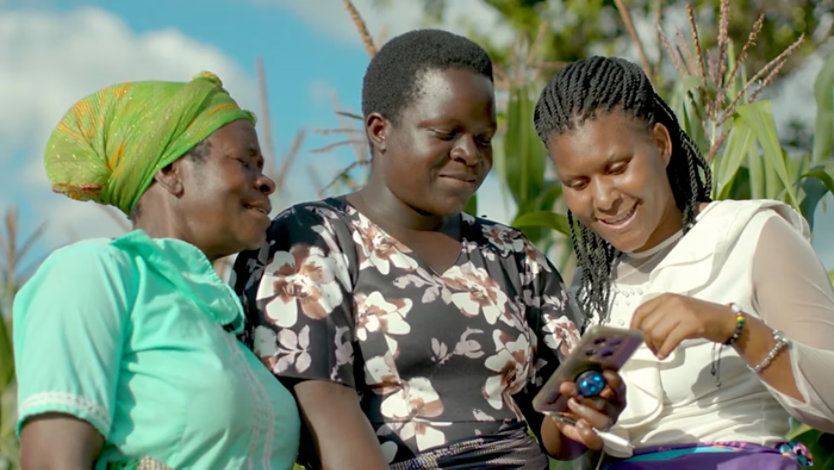 Three women in a rural field in Africa look at a mobile phone together.