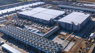 Large industrial data center campus viewed from above, with warehouse-style buildings, cooling infrastructure, and construction equipment surrounding the site.