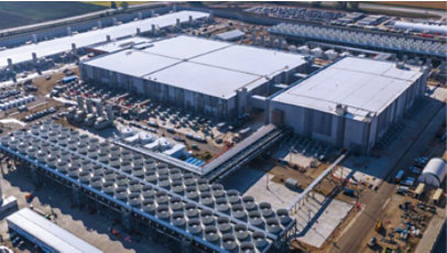 Large industrial data center campus viewed from above, with warehouse-style buildings, cooling infrastructure, and construction equipment surrounding the site.