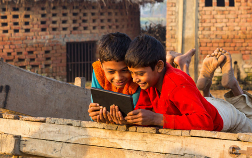 Two children lying on a wooden cart outdoors, smiling while looking at a tablet in a rural village setting.
