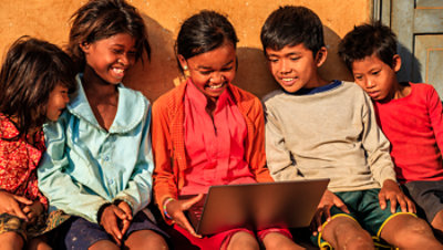 A group of smiling children leaning against a wall outside and viewing a single laptop together.