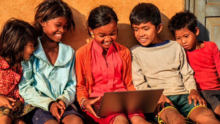 A group of smiling children leaning against a wall outside and viewing a single laptop together.