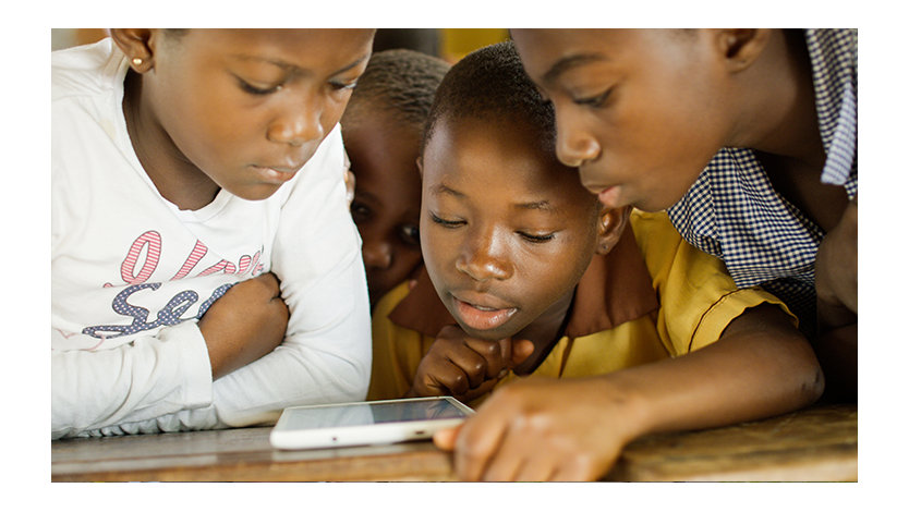 Four young children in Ghana crowded together, looking at one tablet device on a table.