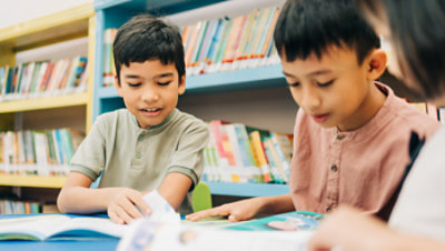 Three children reading books together at a table in a library, with shelves of books in the background.