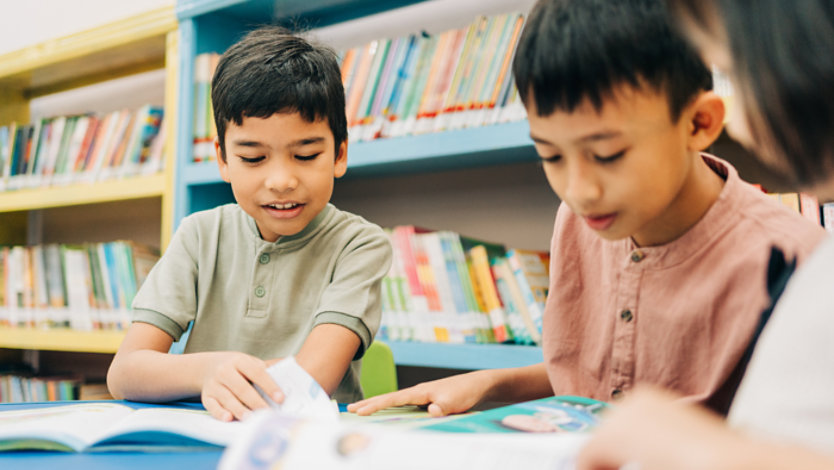 Three children reading books together at a table in a library, with shelves of books in the background.
