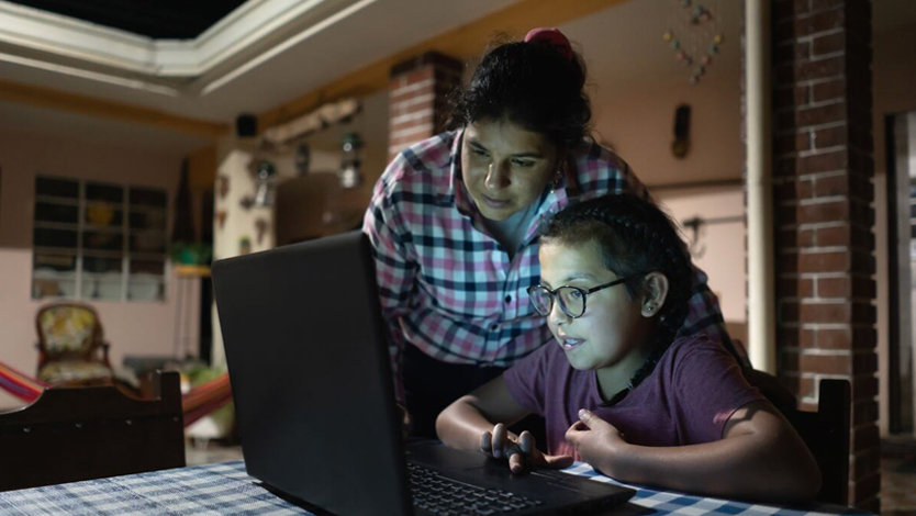 A young girl sits at a dining table looking at a laptop while her mom stands behind her looking over her shoulder.