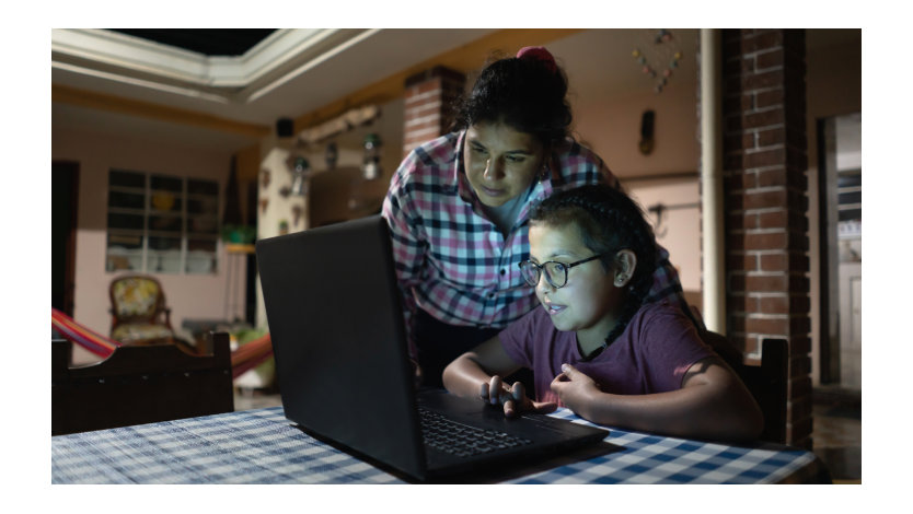 A young girl sits at a dining table looking at a laptop while her mom stands behind her looking over her shoulder.