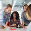 A diverse group of businesswomen look at a digital tablet and discuss a project in their modern office.