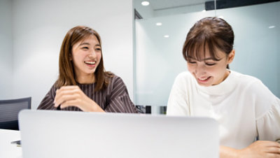 Two women in a business environment sitting in front of a laptop computer smiling and talking.