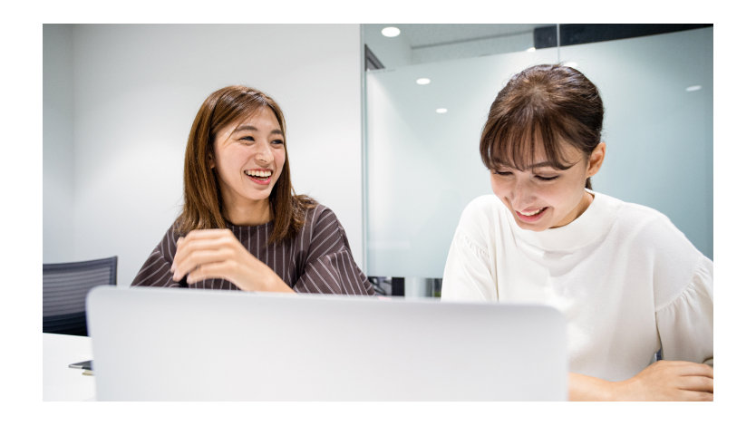 Two women in a business environment sitting in front of a laptop computer smiling and talking.
