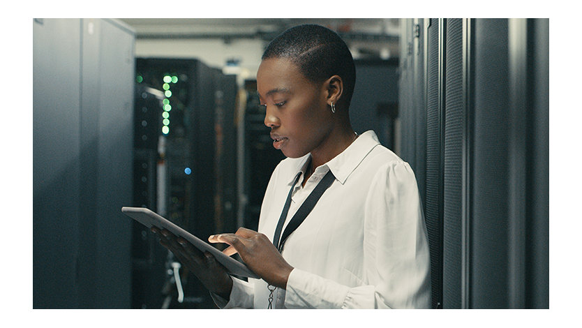 A woman wearing a lanyard standing in an IT server room typing on a digital tablet.